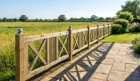 Design Gartenzaun Schloss aus hellem Holz mit Kreuzfüllungen und Edelstahlkugeln an der Terrasse mit Blick auf Wiese und Feld