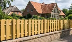 Robuster Zaun mit gerundeten Zaunlatten auf gemauerten Mauersockel als Grundstücksbegrenzung.