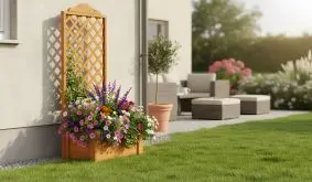 Holzpflanzkasten mit Rankgitter, dicht bepflanzt mit bunten Blumen, steht an einer Hauswand auf einer gepflegten Terrasse mit Garten im Hintergrund.