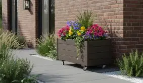 Dunkler Pflanzkasten auf Rollen mit bunten Blumen steht an einer Backsteinwand im Eingangsbereich.