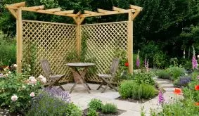 Ruhige Sitzecke mit kleinem Tisch und zwei Stühlen auf einer kleinen Terrasse vor einer Pergola mit Eckverlauf und Rankgittern im Garten.