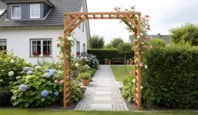 ChatGPT:

Ein schlichter Holz-Rosenbogen rahmt einen gepflegten Gartenweg, umgeben von blühenden Hortensien und Rosen, die eine einladende Atmosphäre schaffen.