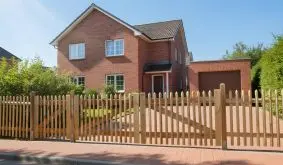 Langer Senkrechtzaun aus naturfarbenem Holz mit Einfahrtstor vor rotem Klinkerhaus mit gepflasterter Einfahrt.