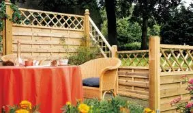 Gemütliche Gartenterrasse mit Holzzaun, Rattanmöbeln und rot gedecktem Tisch, umgeben von bunten Sommerblumen im Vordergrund.
