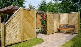 Holz-Sichtschutz im Garten mit diagonaler Lattung, Rosenbogen und Gartenbank, umgeben von blühenden Kletterrosen und Kiesweg.