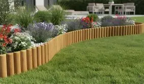 Geschwungene Holz-Rasenkante trennt Rasen von buntem Blumenbeet mit Steinen; im Hintergrund Terrasse mit Gartenmöbeln.