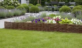 Niedrige Weiden-Beeteinfassung um buntes Blumenbeet im Garten, davor Rasen, im Hintergrund Terrasse mit Sitzgruppe.