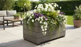 Grauer Holz-Pflanzkübel auf Terrasse, üppig bepflanzt mit weißen und violetten Blüten sowie Farnen im sonnigen Garten.