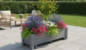 Graues Hochbeet aus Holz, bepflanzt mit bunten Sommerblumen und einem kleinen Strauch, steht auf einer modernen Terrasse im Garten.