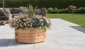 Runder Pflanzkübel aus hellem Holz mit üppigen rosa, weißen und blauen Blüten auf einer modernen Terrassenfläche im Garten.