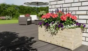 Holzpflanzkasten mit bunten Sommerblumen steht auf einer Terrasse vor einer hellen Hauswand im Garten.