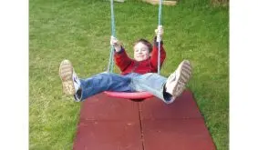 Roter Kunststoff-Schaukelsitz mit blauen Seilen im Garten, Kind schwingt fröhlich darauf mit ausgestreckten Beinen