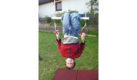 Schaukelringe im Garten: Kind hängt kopfüber an Turnringen mit Polyseilen, trägt rote Jacke und Jeans, spielt im Freien