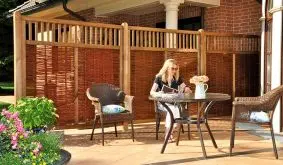 Gemütliche Terrasse mit Sichtschutz aus Holz und Weide, Rattanmöbeln und bunten Blumen. Person liest an rustikalem Gartentisch.