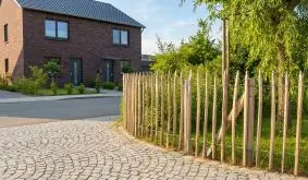 Staketenzaun aus Haselnussholz als natürliche Grundstücksbegrenzung entlang Einfahrt mit modernem Haus, Grünfläche und Bäumen.