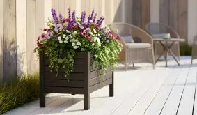 Hoher schwarzer Holzpflanzkasten voller bunter Blumen auf einer sonnigen Terrasse.