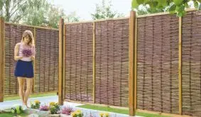 Weidenzaun als Sichtschutz im Garten mit Holzpfosten, davor Blumenbeete mit gelben und violetten Blüten.