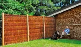 Rustikaler Weidenzaun im Garten neben Holzlager, mit Hund und Person auf Bank sitzend, umgeben von grünen Bäumen.