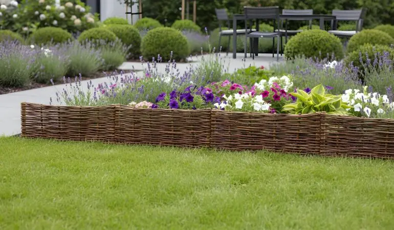 Niedrige Weiden-Beeteinfassung um buntes Blumenbeet im Garten, davor Rasen, im Hintergrund Terrasse mit Sitzgruppe.