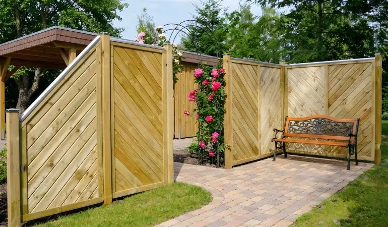 Holz-Sichtschutz im Garten mit diagonaler Lattung, Rosenbogen und Gartenbank, umgeben von blühenden Kletterrosen und Kiesweg.