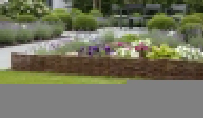 Niedrige Weiden-Beeteinfassung um buntes Blumenbeet im Garten, davor Rasen, im Hintergrund Terrasse mit Sitzgruppe.