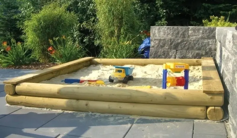 Sandkasten aus Rundholz im Garten Offener Sandkasten aus Rundholz mit Spielzeug auf einer gepflasterten Fläche im Garten