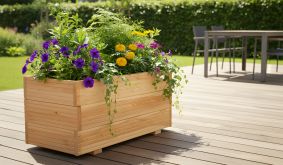 Holzpflanzkasten mit bunten Sommerblumen auf einer sonnigen Terrasse, umgeben von Gartenmöbeln und grünem Rasen.
