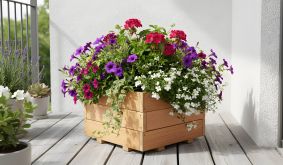 Holzpflanzkasten mit bunten Blumen auf hellem Balkonboden nahe einer weißen Wand.